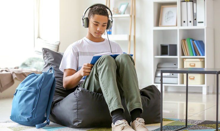 teenager sitting on beanbag chair, reading book with headphone on